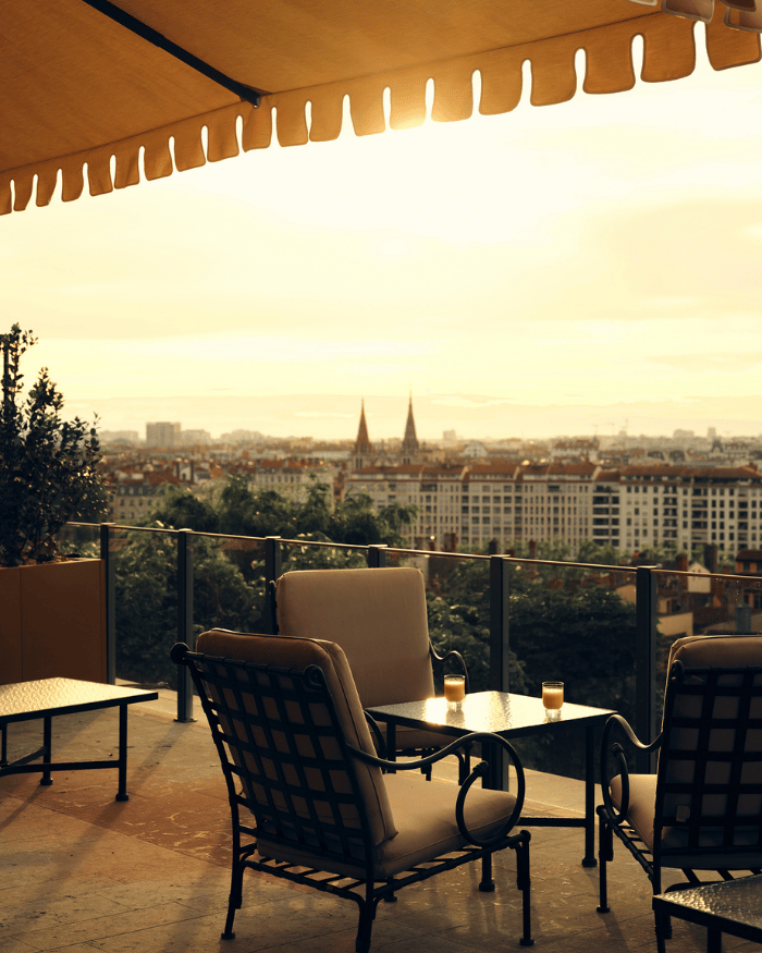 Outdoor patio with cushioned chairs and a table overlooking cityscape at sunset under a canopy.