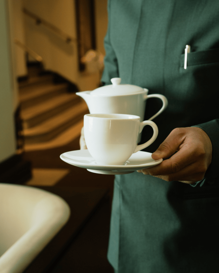 Person in green coat holding a white teapot and a teacup on a saucer indoors.