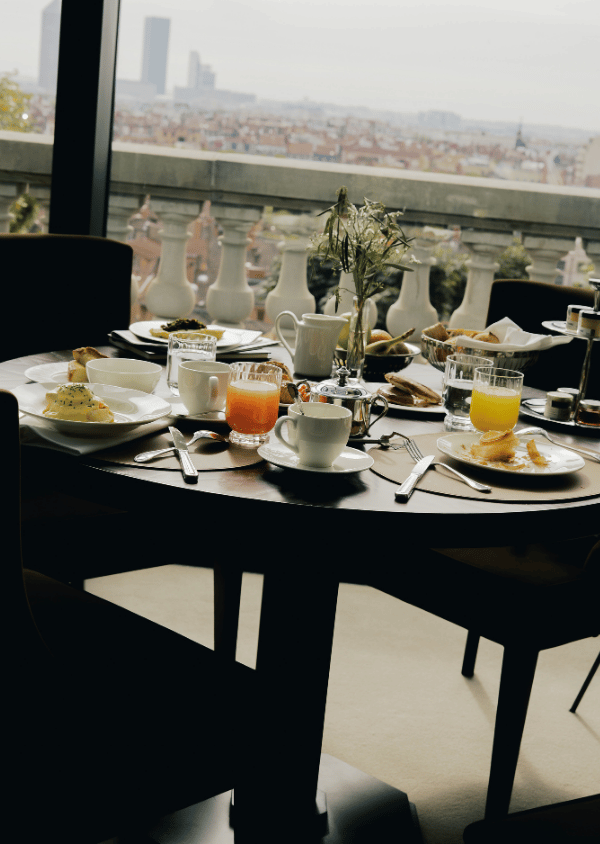 Round table with breakfast items like juice, coffee, eggs, and pastries near a window overlooking city buildings.