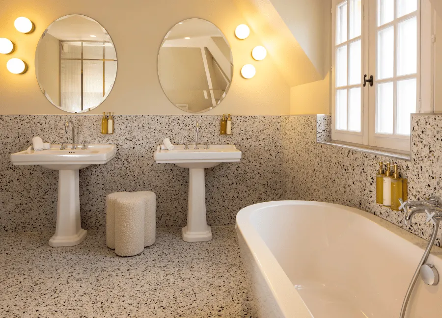 Bagno luminoso dal design retrò, con due lavabi a colonna, specchi rotondi circondati da applique e pavimento in terrazzo.