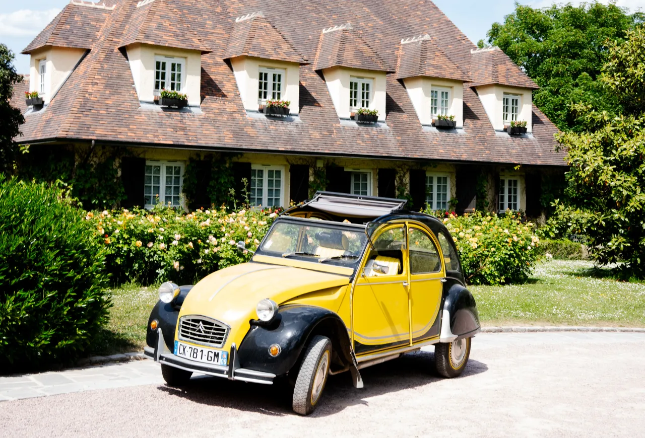 Yellow Citroën 2CV parked in front of a large Norman-style house surrounded by rosebushes.