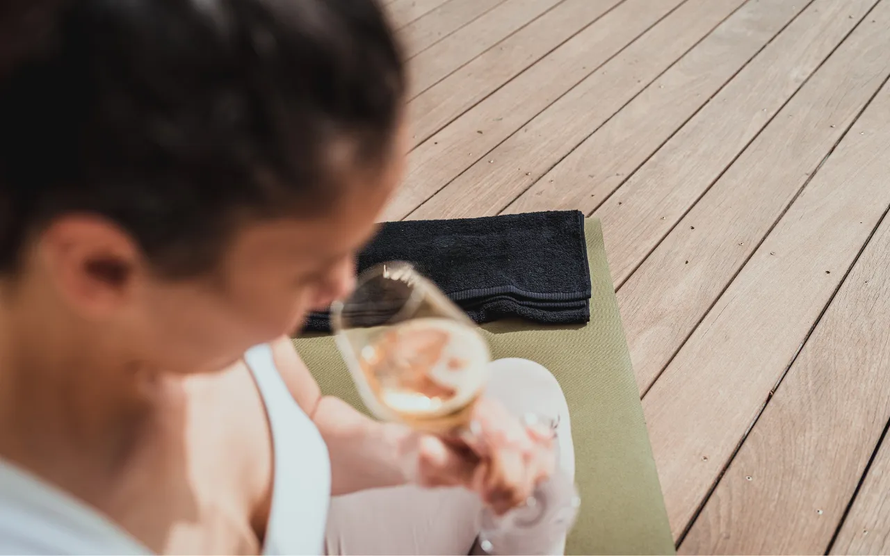 Person sitting on a yoga mat holding a glass of champagne on a wooden deck.