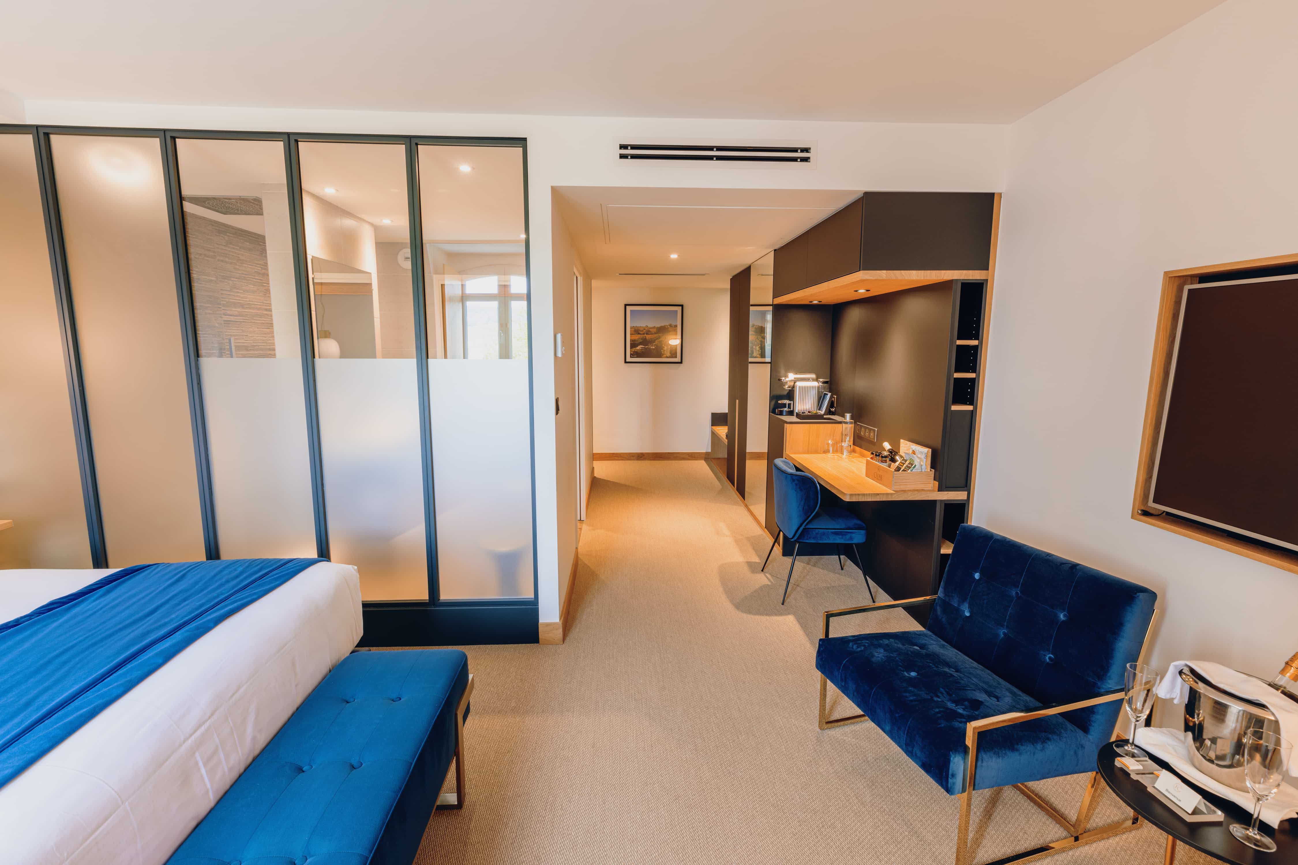 Eine moderne Hotelsuite mit einem Bett mit blauer Tagesdecke, einer Sitzecke mit blauen Samtsesseln und Blick auf den Flur, der zum Badezimmer führt.