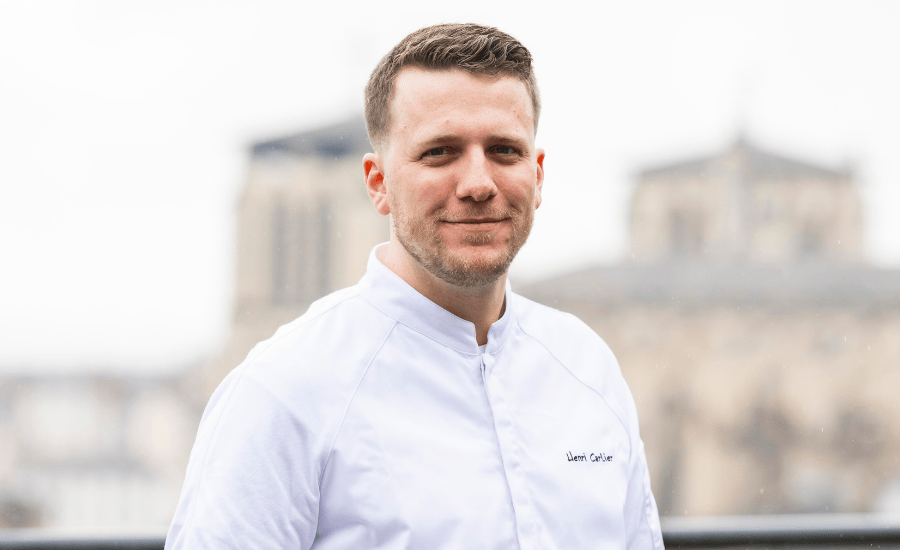 Ritratto dello chef Henri Carlier in giacca bianca con Notre-Dame de Paris su sfondo sfocato.