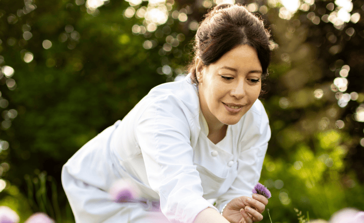 Lo chef Nawal Rezagui sorride mentre raccoglie fiori commestibili in un giardino soleggiato.