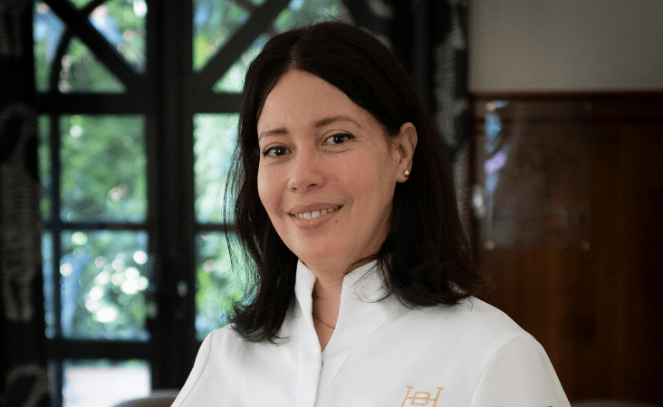 Ritratto dello chef Nawal Rezagui in giacca da cucina bianca, sorridente davanti a una tettoia di legno.