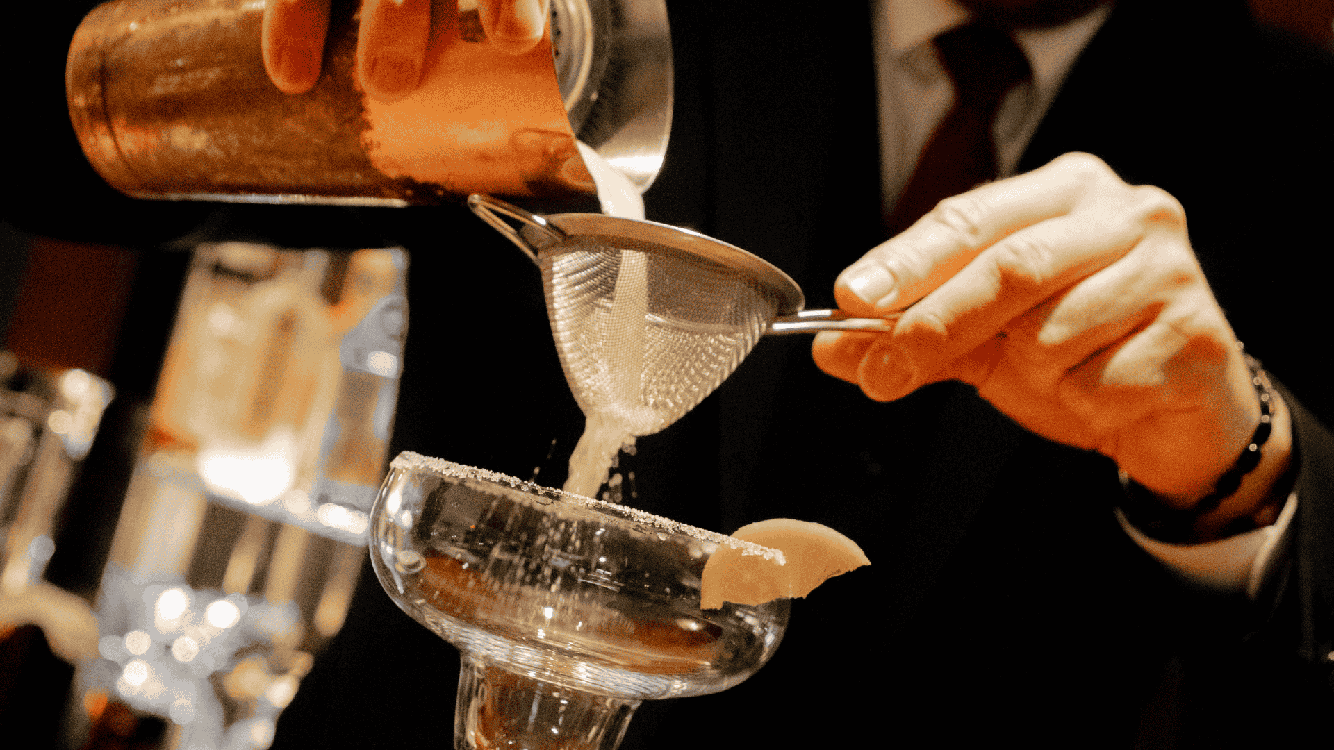 A bartender in a suit pouring a cocktail from a shaker through a fine strainer into a frosted glass.