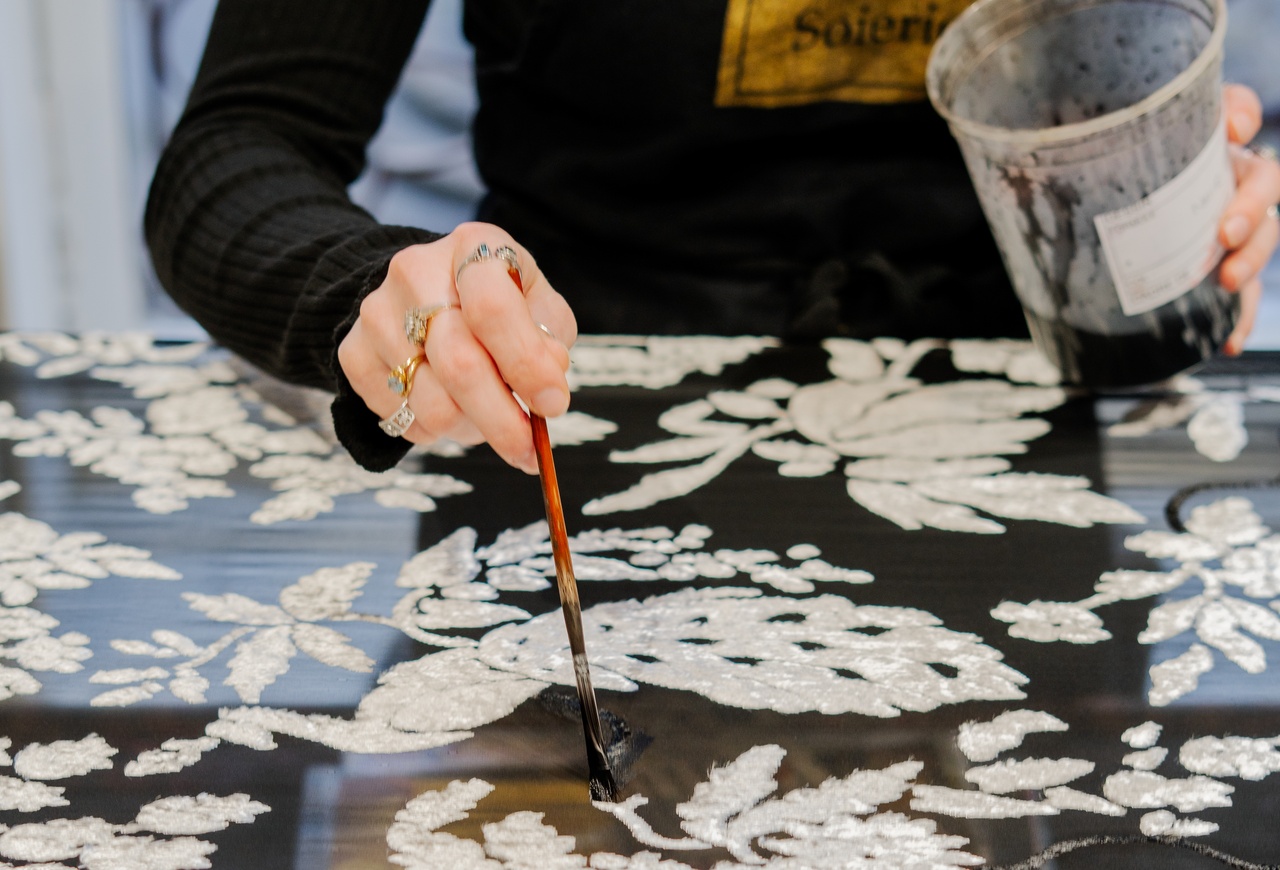 Close-up of a hand applying black dye with a brush to a fabric embroidered with white floral patterns, in a silk workshop. Villa Florentine, Lyon.