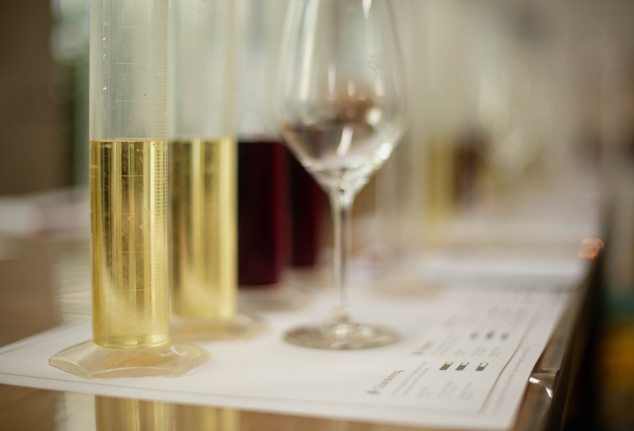 Test tubes containing white wine samples, with a tasting glass and red wine bottles in the background, during an oenological analysis. Villa Florentine, Lyon