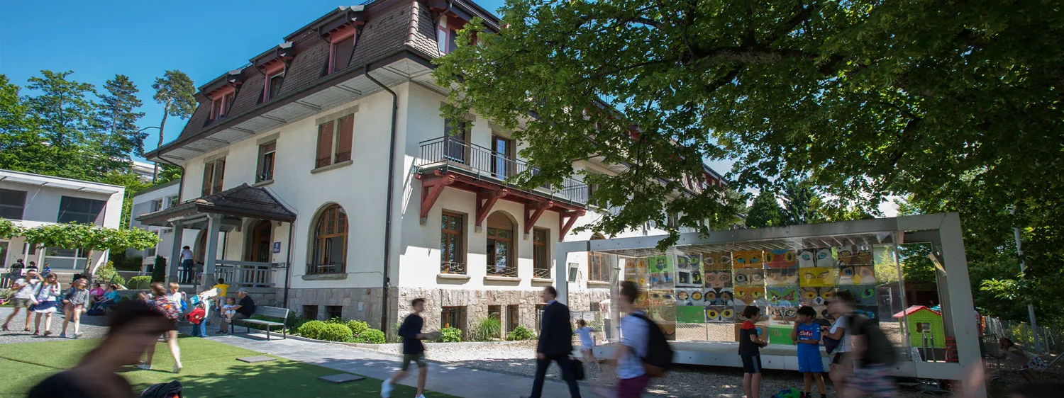 Schoolyard scene with children playing and walking near a large white multi-story building surrounded by trees and a colorful art display.