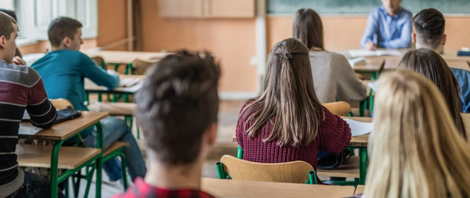 Students sitting at desks facing a teacher who is seated at a desk in a classroom.