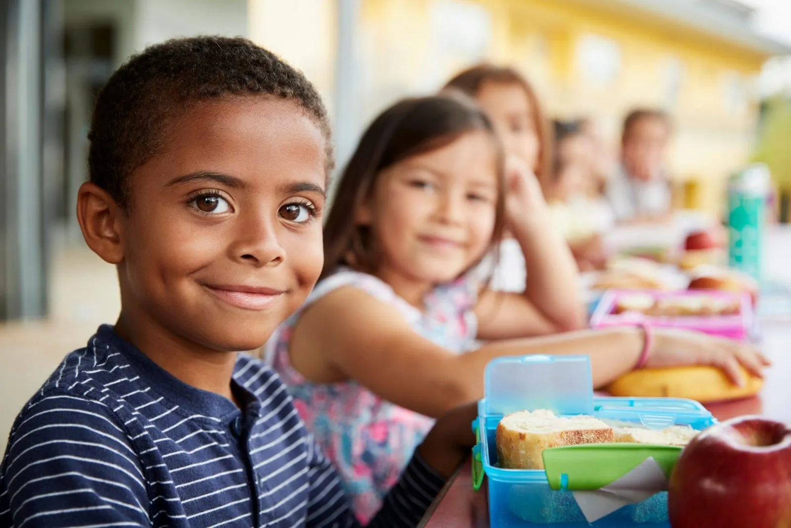 Smiling boy sitting at a table with a lunchbox containing bread and an apple, with other children blurred in the background.