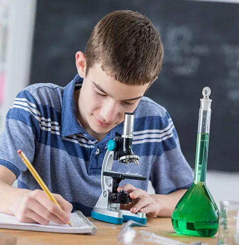Student in a blue striped shirt using a microscope and taking notes at a desk with green liquid in a flask.