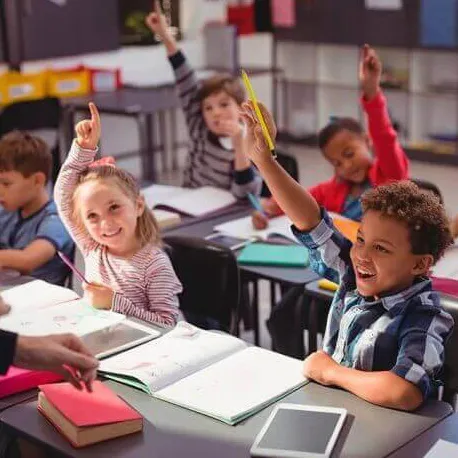 Young diverse students sitting at desks in a classroom eagerly raising their hands to answer a question.