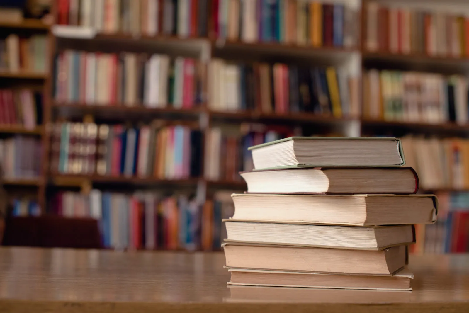 Stack of six hardcover books on a wooden table with blurred bookshelves in the background.