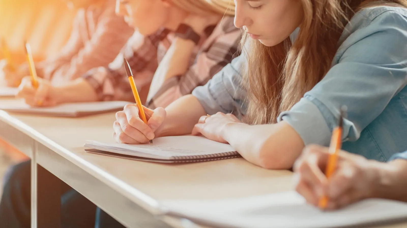 Students sitting at desks writing with pens in notebooks during a classroom setting.