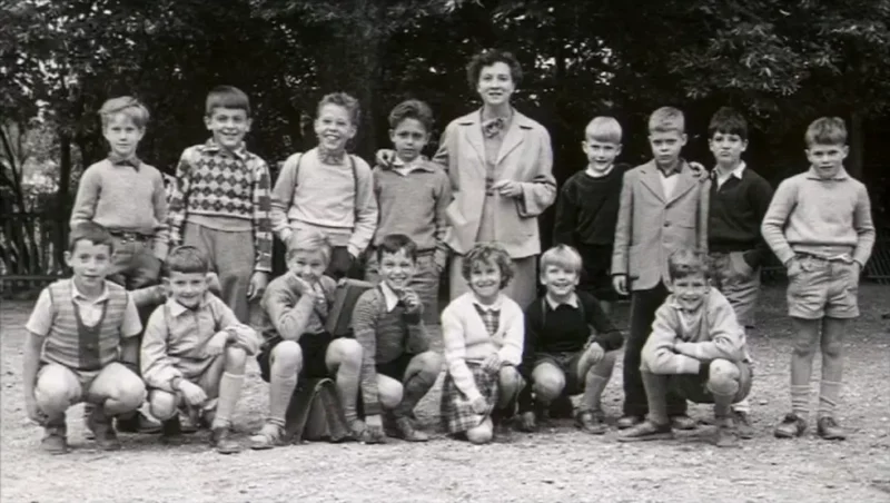 Black and white photo of a group of 16 children and one adult woman posing outdoors with trees in the background.