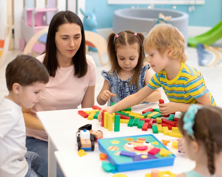 Four children and a woman sitting around a table playing with colorful wooden blocks and educational toys in a bright playroom.