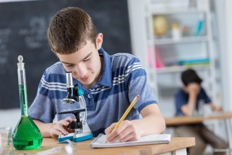 A student in a blue striped shirt looking through a microscope and taking notes in a classroom with chemistry equipment on the desk.