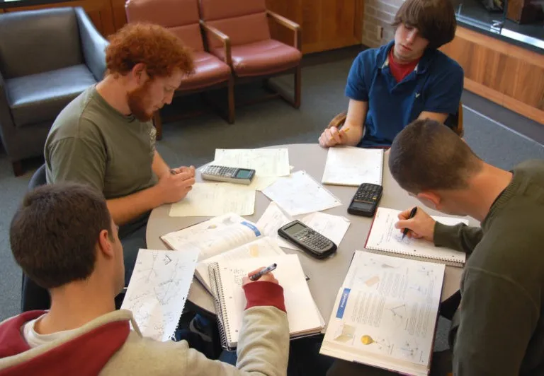Four young men sitting around a table studying with notebooks, textbooks, calculators, and scattered papers.