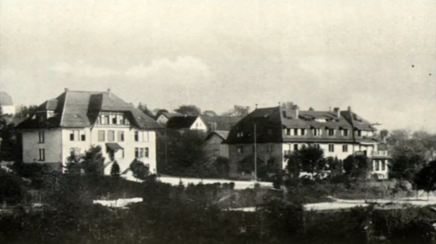 Black and white photo of two large, early 20th-century European-style buildings surrounded by trees and a road in the foreground.