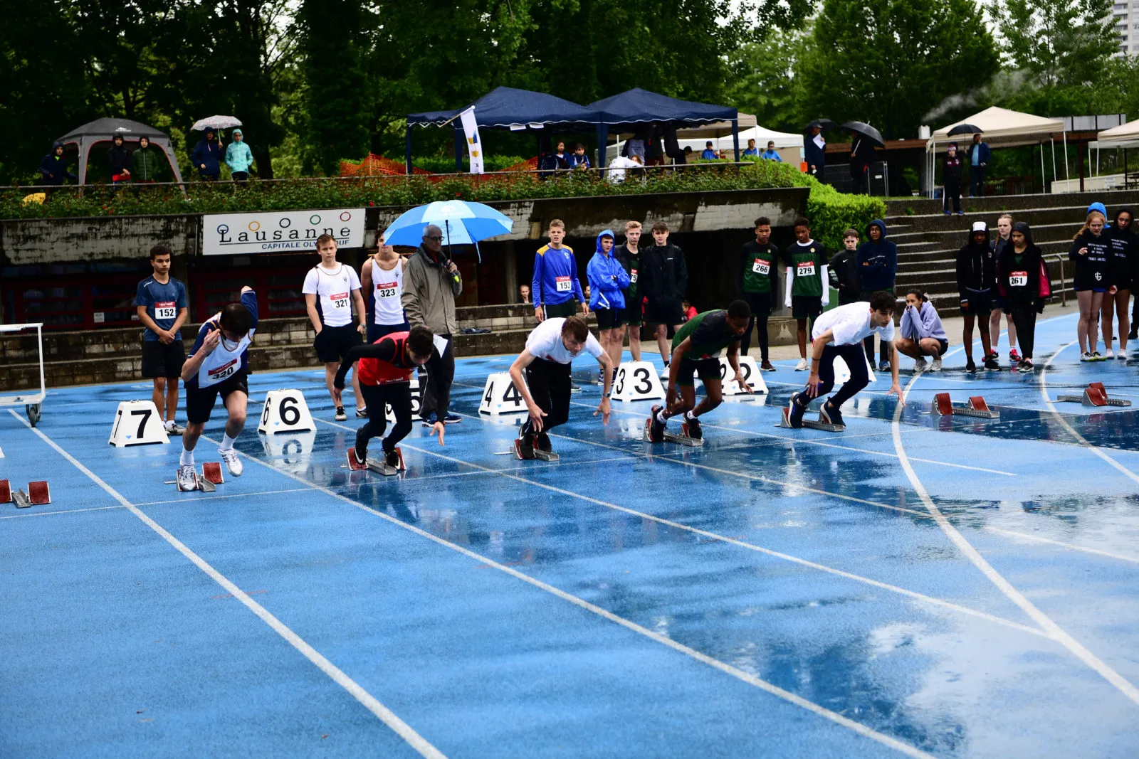Young male athletes poised at the starting blocks on a wet blue track preparing to sprint, with spectators and officials standing in the background under umbrellas and tents.