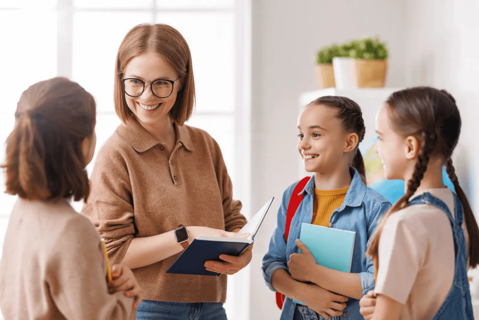 Smiling teacher holding an open book while talking to three happy schoolgirls in a bright classroom.