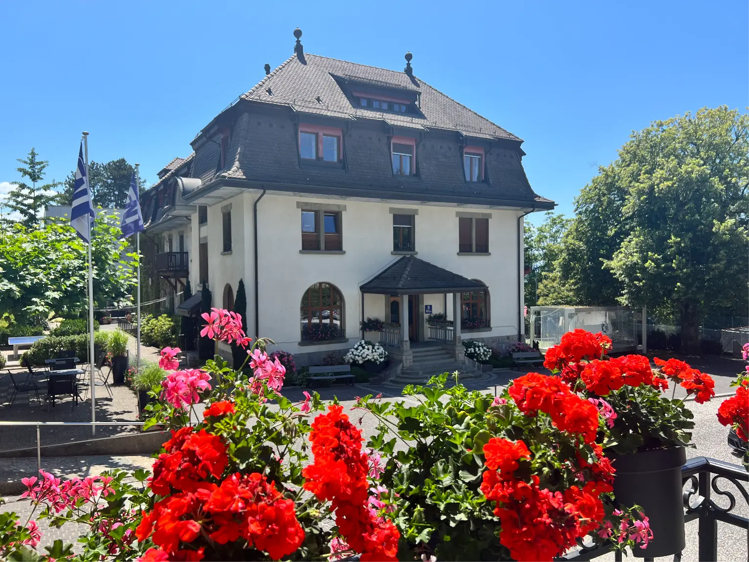 Historic white three-story building with a dark roof, surrounded by bright red and pink flowers and green trees under a clear blue sky.