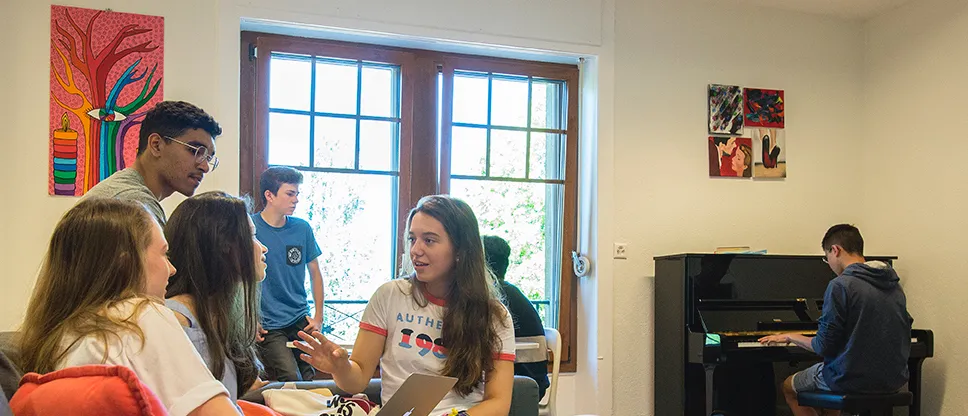 A group of young people sitting and standing near a window talking, with one person playing a piano in the background.