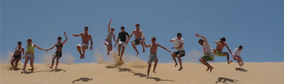 Group of young people jumping off a sand dune under a clear blue sky.