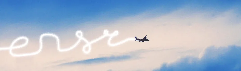 Airplane flying in a partly cloudy sky leaving a white smoke trail forming cursive letters.