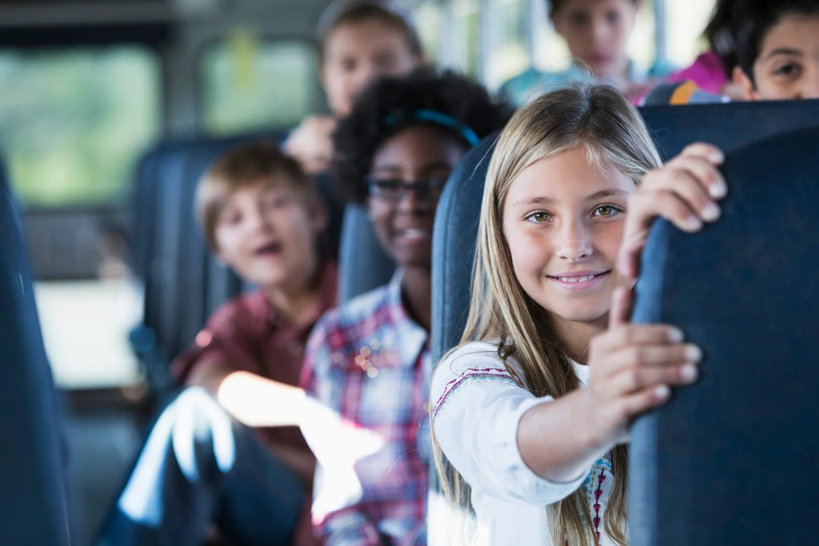 Smiling diverse schoolchildren seated on a bus, with a blonde girl in the foreground holding the seat in front.