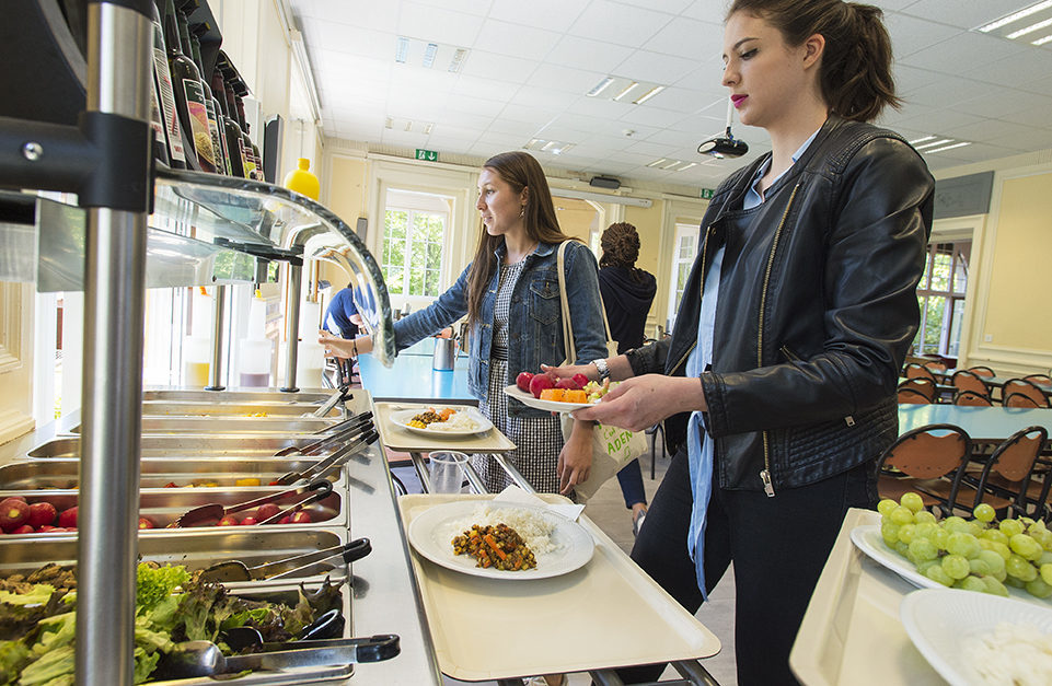 Two women serving themselves food from a cafeteria buffet line; one holds a plate with fruit, the other reaches for food.
