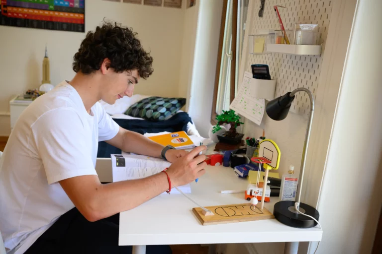 Teenage boy sitting at a white desk in a bedroom, interacting with a small basketball hoop game, with study materials and a desk lamp nearby.