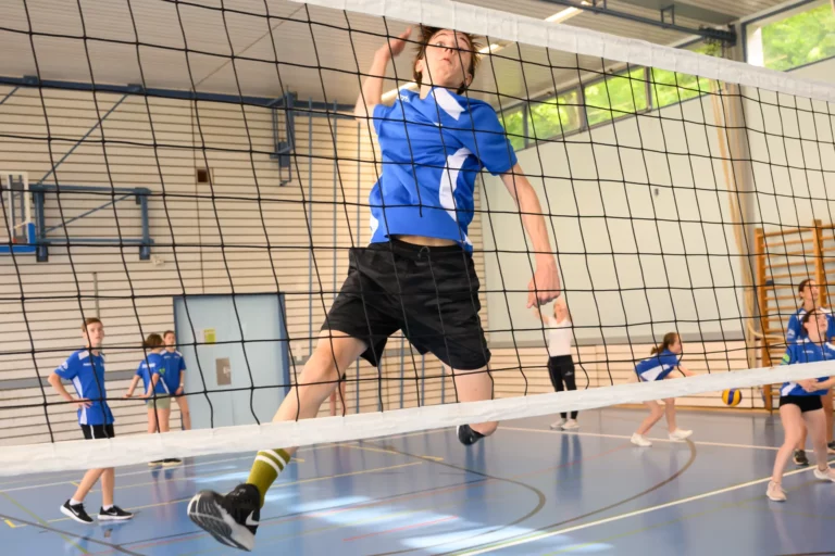 Teenage boy in blue sports jersey jumping high near a volleyball net in an indoor gym with other players watching.