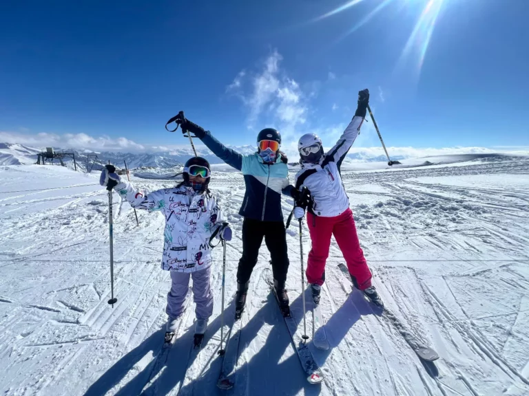 Three skiers posing with ski poles raised on a sunny snowy mountain slope under a blue sky.