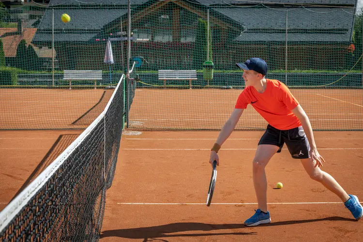 Boy in orange shirt and black shorts playing tennis on an outdoor clay court, reaching to hit a ball near the net.