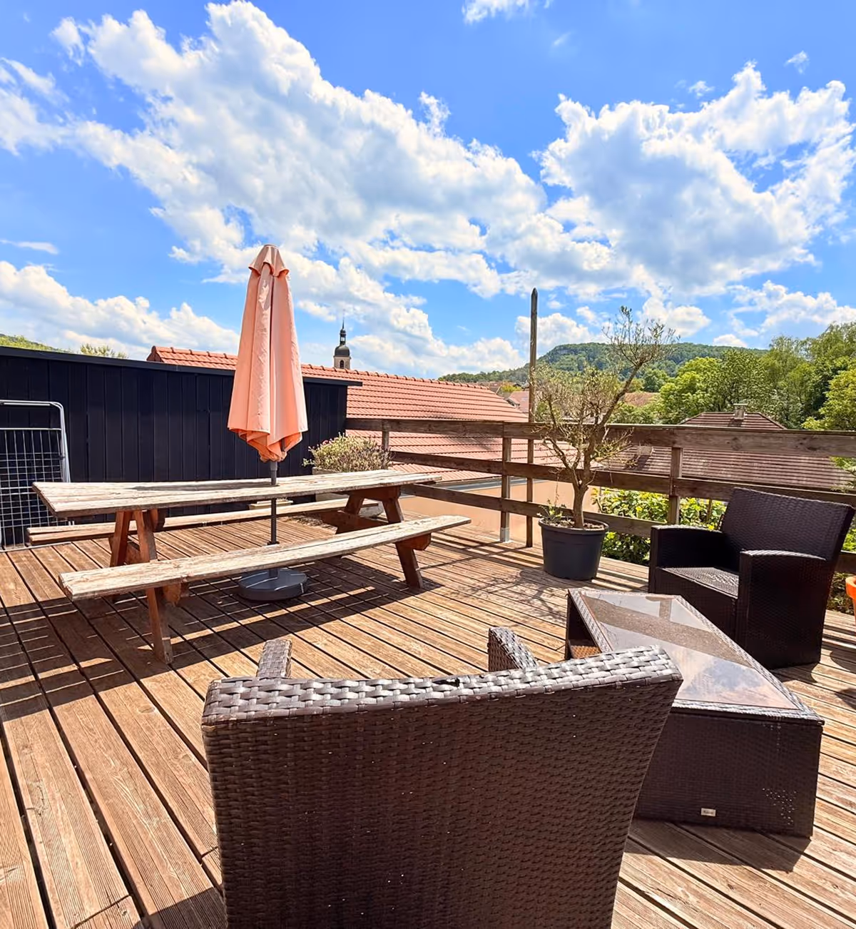 Terrasse en bois avec chaises en osier foncé, table basse en verre, table de pique-nique en bois, parasol fermé et plante en pot sous un ciel bleu partiellement nuageux.