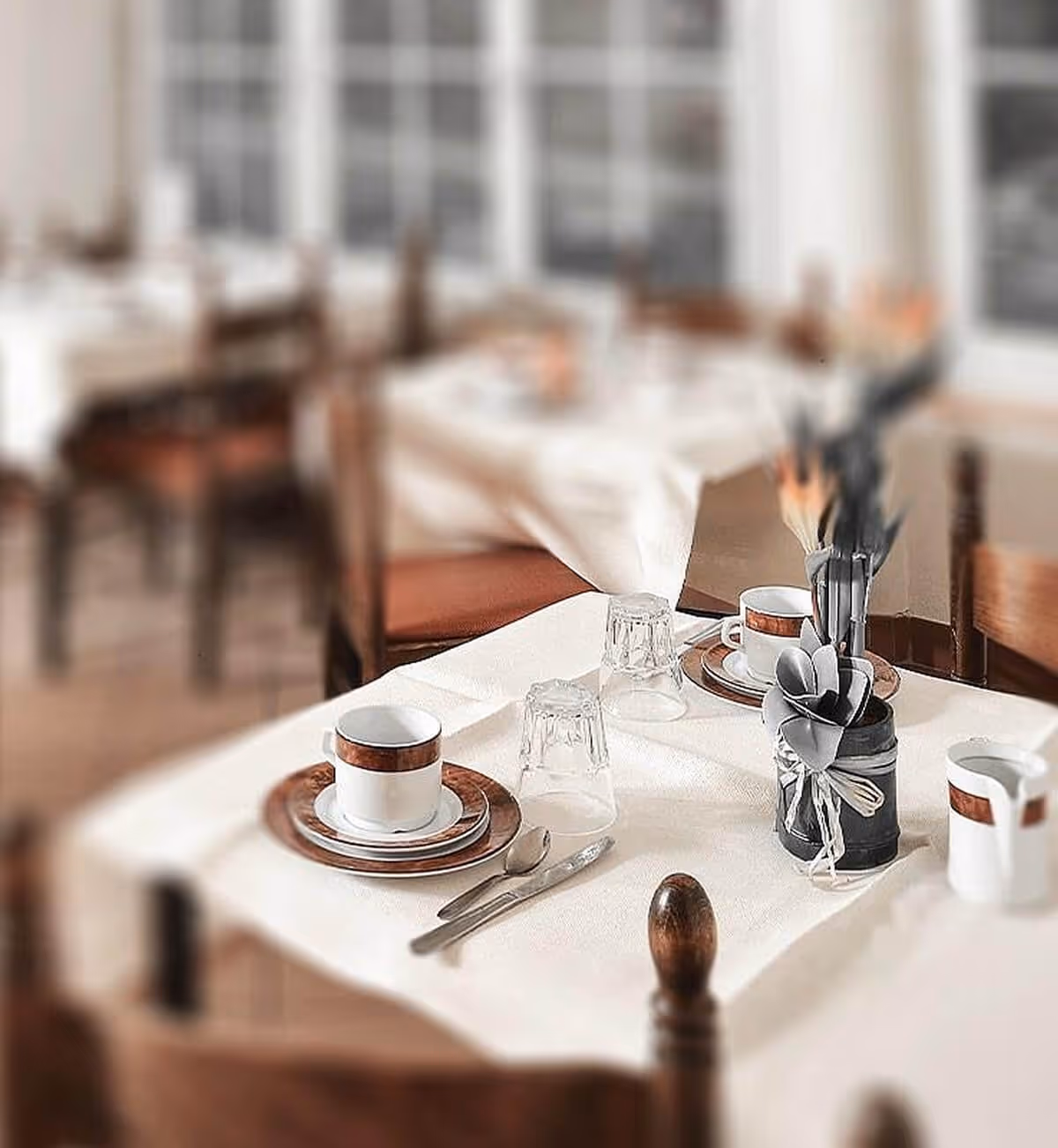 Dans une salle à manger à l'éclairage tamisé, une table est dressée pour deux avec une nappe blanche, des tasses, des soucoupes, des verres, des couverts et un petit pot décoratif contenant ustensiles et serviettes.