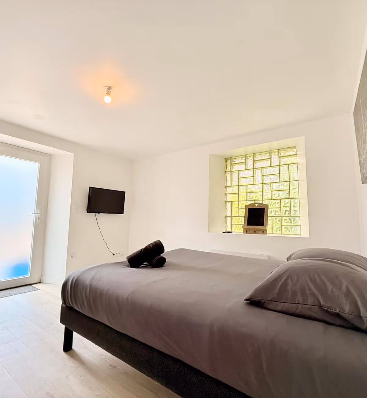 Chambre minimaliste avec un lit gris, des serviettes roulées sur le lit, une télévision fixée au mur et une fenêtre en pavés de verre avec un petit miroir.