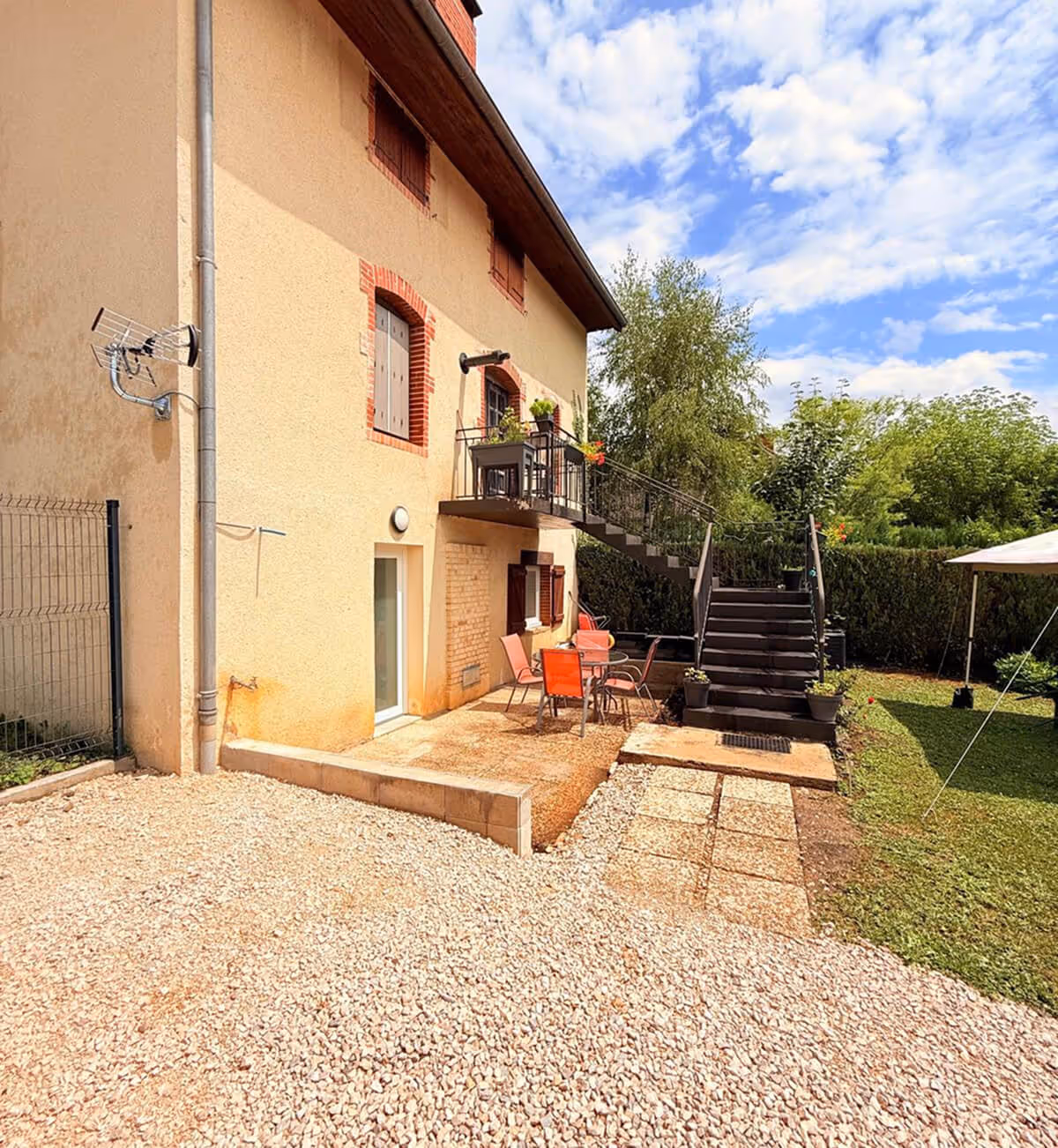 Terrasse extérieure avec chaises et table orange à côté d'une maison beige à deux étages, escalier menant à un balcon avec des plantes en pot et pelouse verte sous un ciel partiellement nuageux.