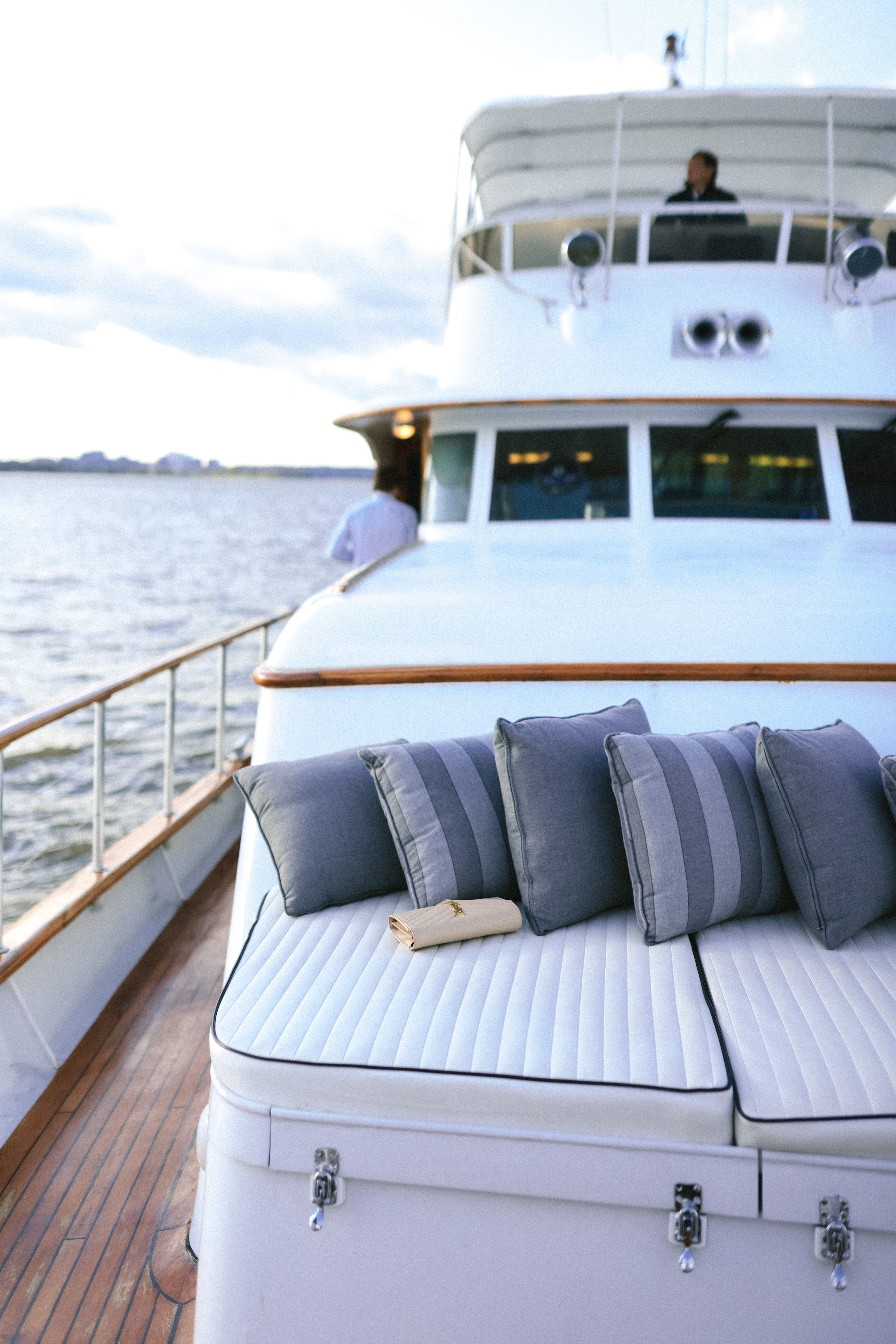 White yacht deck with cushioned seating and blue-gray pillows beside calm water under a cloudy sky.