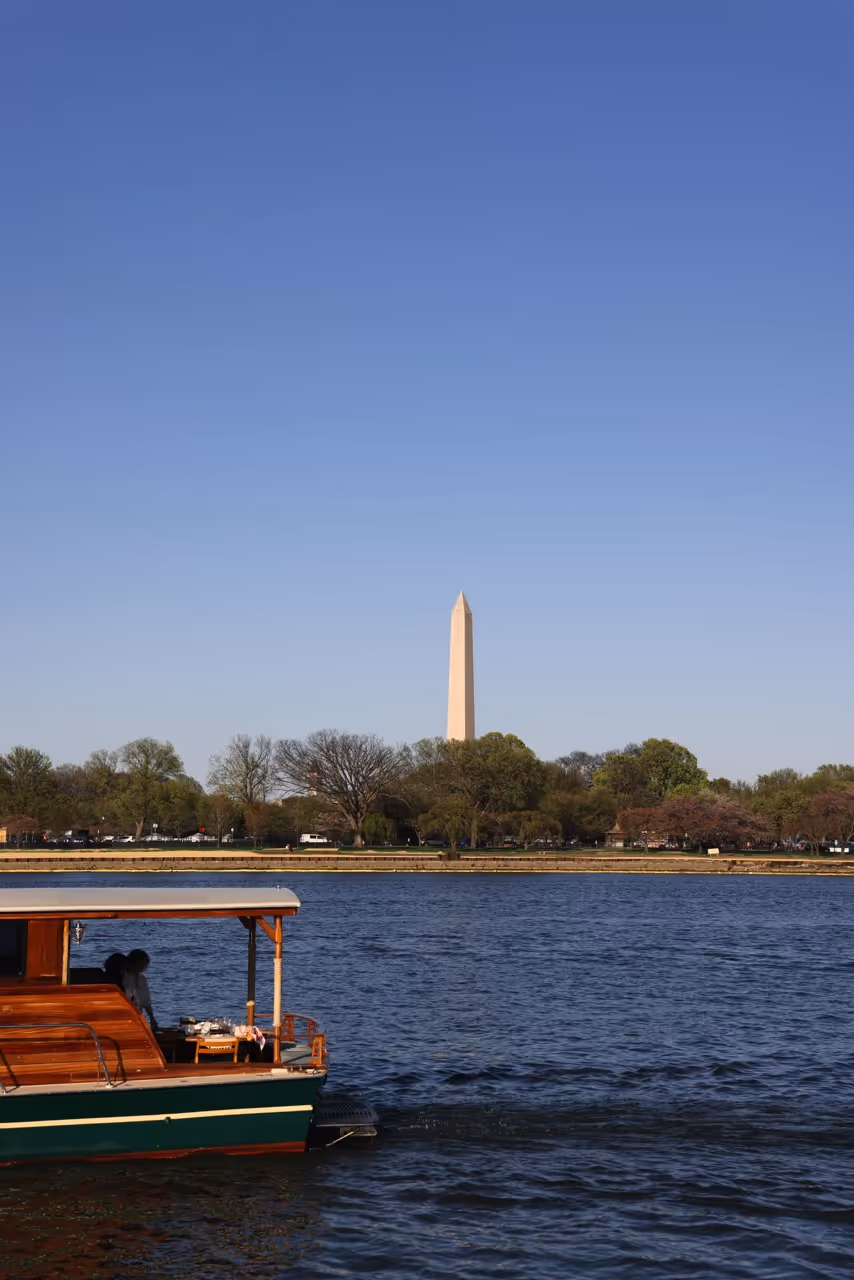 Vintage wooden yacht on water with the Washington Monument and trees in the background under a clear blue sky.
