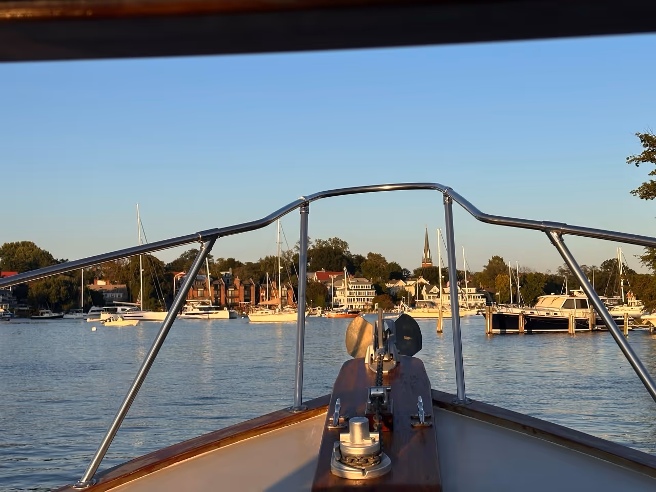 View from a boat's bow with metal railing looking out over water towards a marina with docked boats and waterfront houses under a clear sky.