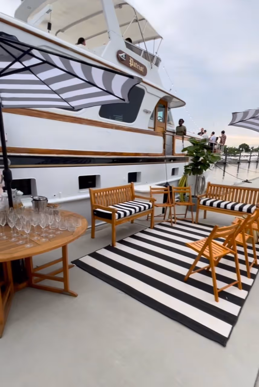 Outdoor lounge area with wooden chairs, striped cushions, a matching striped rug, and umbrellas beside a large white yacht named Patriot docked at a marina.