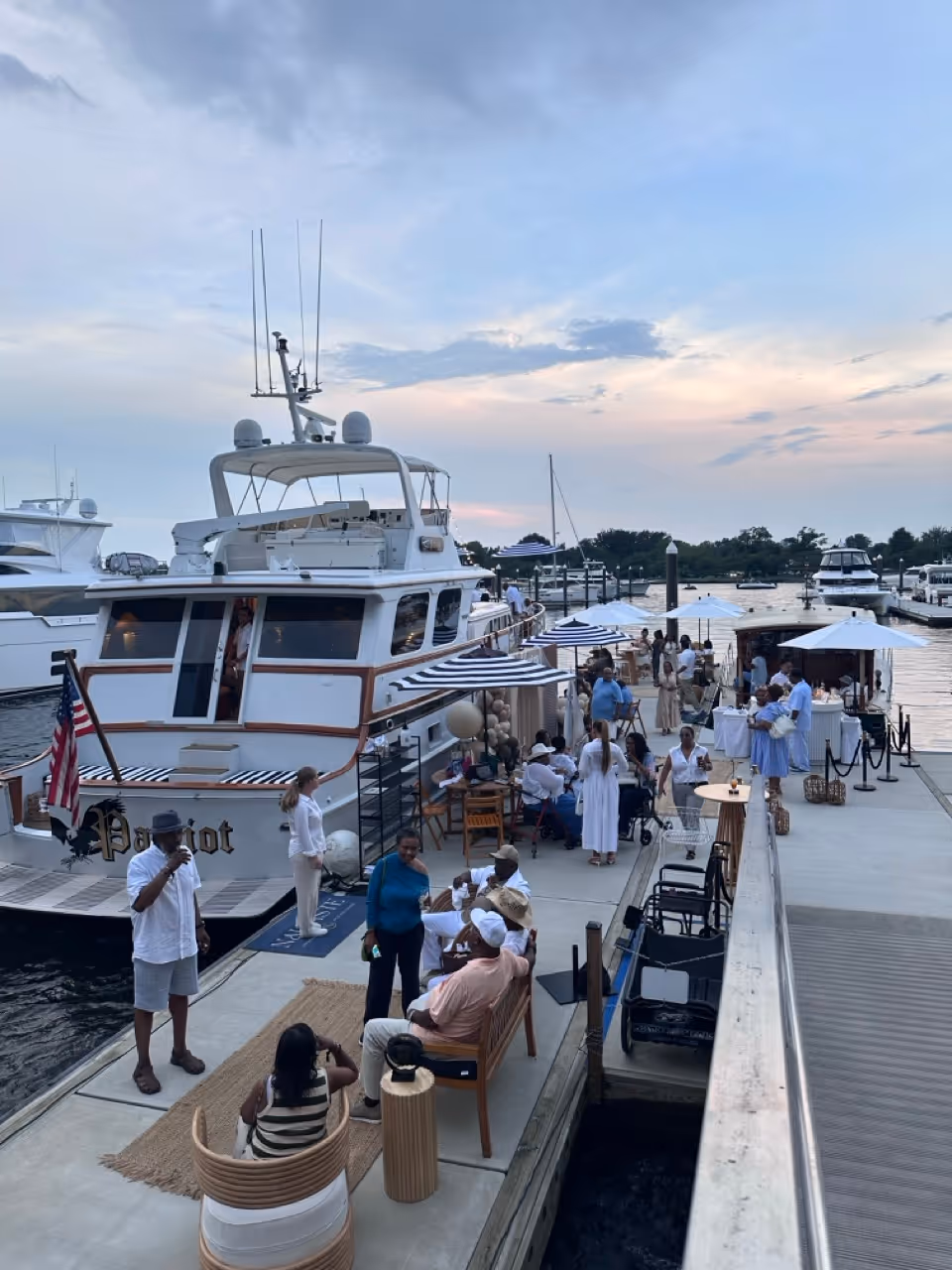 People socializing on a dock beside a white yacht named Patriot during dusk with umbrellas and seating arrangements.
