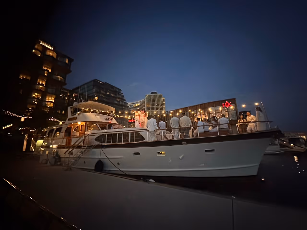 Group of people dressed in white socializing on a docked yacht at dusk with illuminated buildings and string lights in the background.