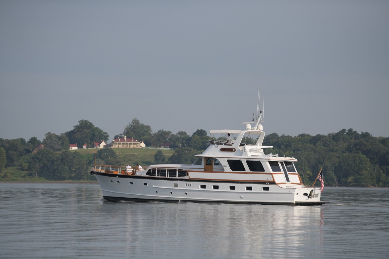 White luxury motor yacht named Patriot cruising on calm water with a wooded shoreline and historic building in the background.