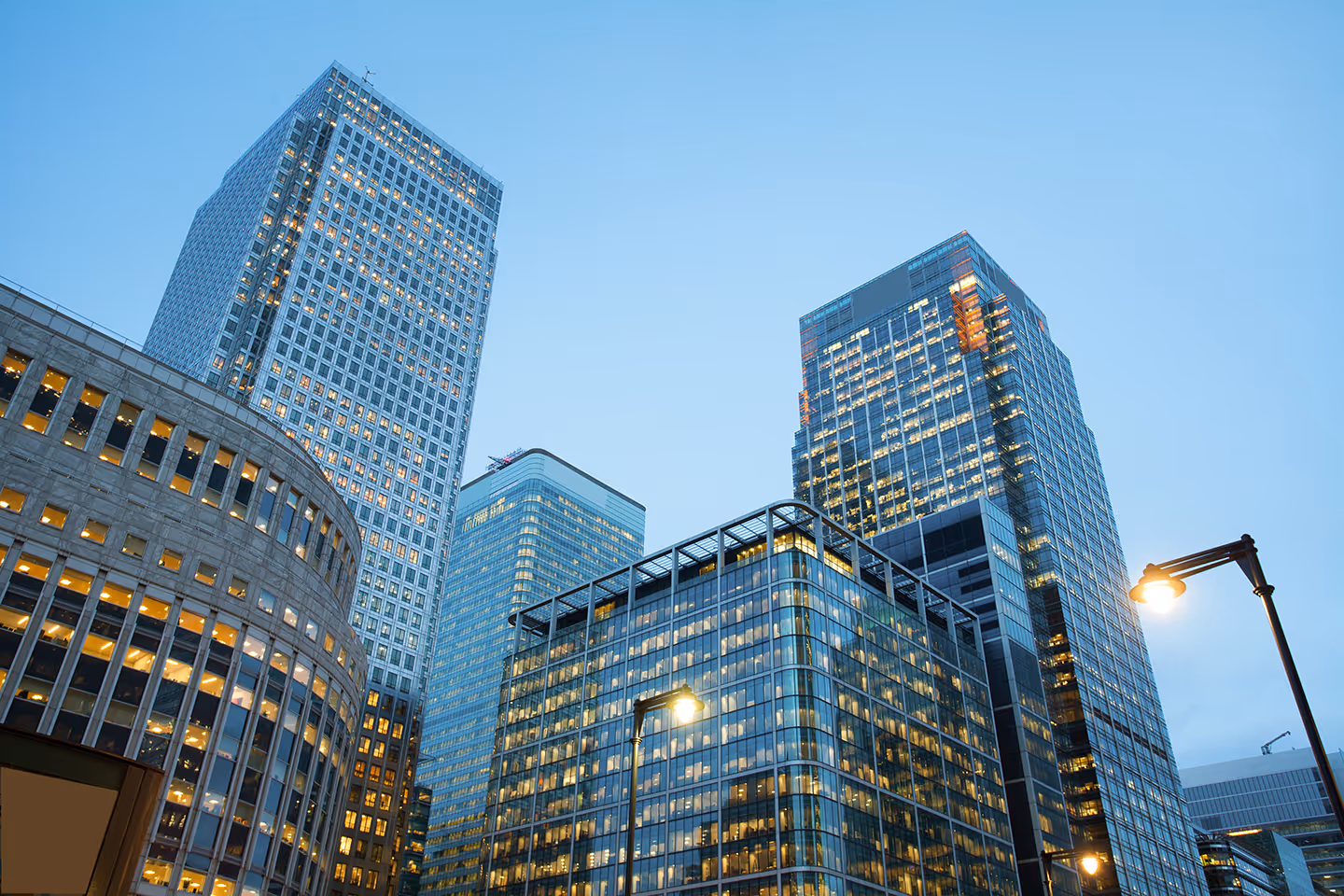Evening view of illuminated modern glass and concrete skyscrapers in London's Canary Wharf with street lamps.