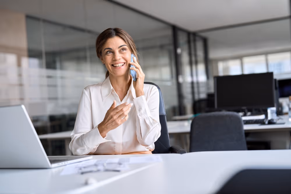 Smiling businesswoman in white shirt talking on a smartphone at a desk in a modern office.